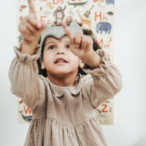 A child in a dress points towards an alphabet poster, showing curiosity and learning.