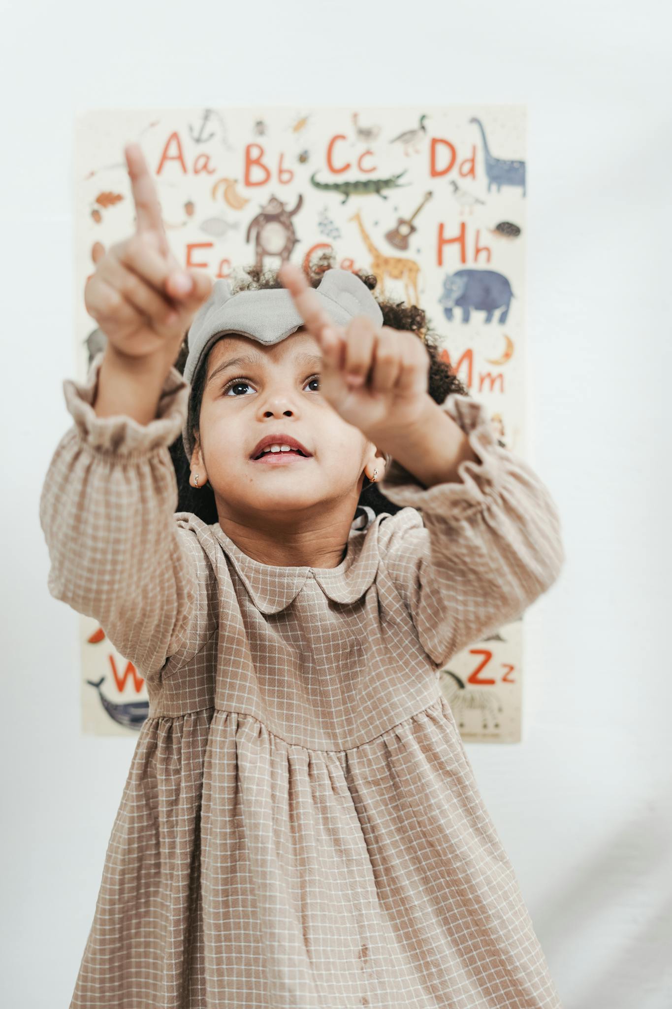 A child in a dress points towards an alphabet poster, showing curiosity and learning.
